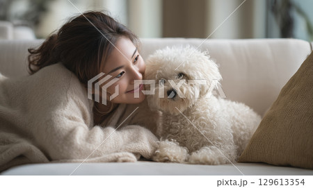 Woman cuddling with her fluffy white dog on a cozy sofa, sharing a warm smile 129613354
