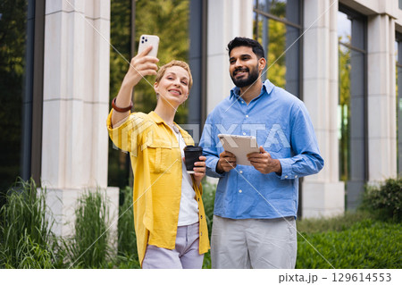 A diverse couple taking a selfie outdoors with a smartphone and tablet in front of a modern building. 129614553