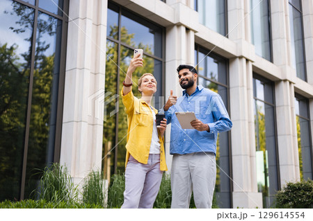 A diverse pair poses outside a modern building, one taking a selfie while the other holds a tablet, both displaying positive gestures. 129614554