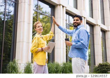 A diverse pair celebrating a success outdoors in front of a modern building, high-fiving with smiles and holding a tablet and coffee. A diverse pair celebrating a success outdoors in front of a modern building, high-fiving with smiles and holding a tablet and coffee. 129614557