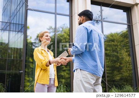 A business handshake between two people in front of a modern building. The woman holds a tablet, and the man holds a coffee cup. A business handshake between two people in front of a modern building. The woman holds a tablet, and the man holds a coffee cup. 129614577
