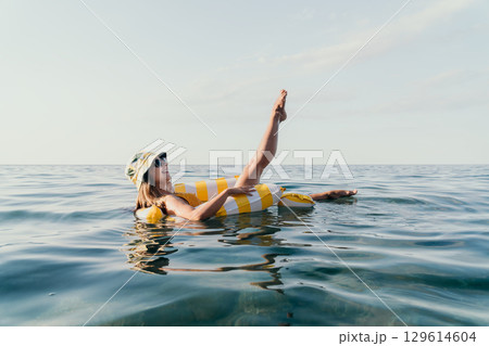 Woman Float Ring Ocean - A woman relaxes in the ocean on a yellow and white striped float ring while wearing a sun hat. 129614604