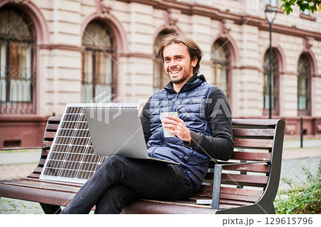 Man sits on wooden bench in urban setting, working on laptop with cup of coffee in hand. Solar panel rests beside, emphasizing sustainable, mobile workspace amidst backdrop of historic architecture. Man sits on wooden bench in urban setting, working on laptop with cup of coffee in hand. Solar panel rests beside, emphasizing sustainable, mobile workspace amidst backdrop of historic architecture. 129615796