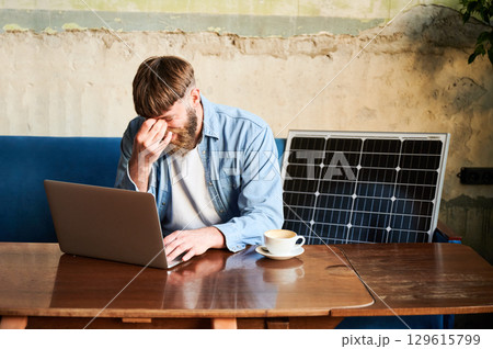 Bearded man works on laptop at wooden table, with cup of coffee. Solar panel nearby, emphasizing sustainable eco-friendly workspace that combines technology and sustainability in cozy environment. 129615799