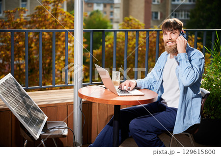 Man works on laptop and smartphone at outdoor table, with glass of iced coffee. Solar panel nearby, emphasizing sustainable, modern eco-friendly workspace that combines technology and sustainability. Man works on laptop and smartphone at outdoor table, with glass of iced coffee. Solar panel nearby, emphasizing sustainable, modern eco-friendly workspace that combines technology and sustainability. 129615800