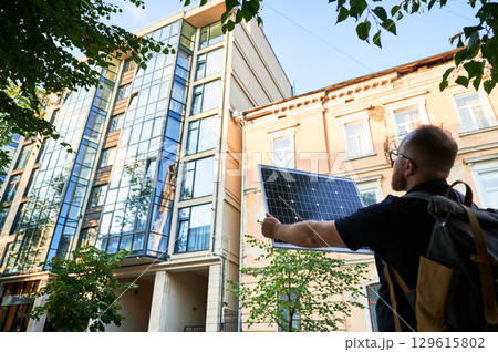Man holding photovoltaic solar panel in front of new building. Concept of integration of sustainable renewable energy sources into modern architecture. Man holding photovoltaic solar panel in front of new building. Concept of integration of sustainable renewable energy sources into modern architecture. 129615802