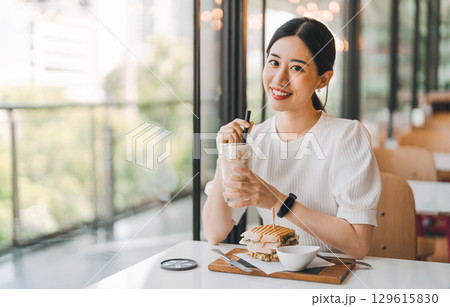 Happy asian woman healthy eating breakfast sandwich at cafe sitting near window indoors on day Happy asian woman healthy eating breakfast sandwich at cafe sitting near window indoors on day 129615830