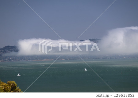 青空と白い帆のヨットの海と霧のような雲のかかるサンフランシスコの街並みの陸の見える風景 129615852