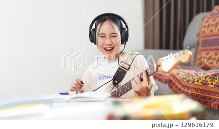 Young adult asian woman playing electric guitar music wearing headphones enjoy hobby at home 129616179