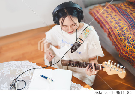 Young adult asian woman playing electric guitar music wearing headphones enjoy hobby at home 129616180