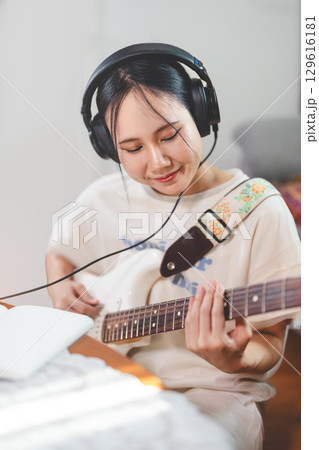 Young adult asian woman playing electric guitar music wearing headphones enjoy hobby at home Young adult asian woman playing electric guitar music wearing headphones enjoy hobby at home 129616181