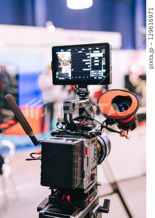 Professional digital video camera with external monitor and accessories, mounted on a tripod. The camera is equipped with a lens and an external battery. Film production Professional digital video camera with external monitor and accessories, mounted on a tripod. The camera is equipped with a lens and an external battery. Film production 129616971