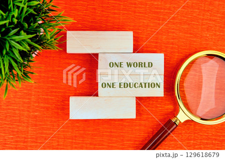 Concept words ONE WORLD ONE EDUCATION assembled from wooden blocks in composition with magnifying glass and green plant on orange background, top view Concept words ONE WORLD ONE EDUCATION assembled from wooden blocks in composition with magnifying glass and green plant on orange background, top view 129618679