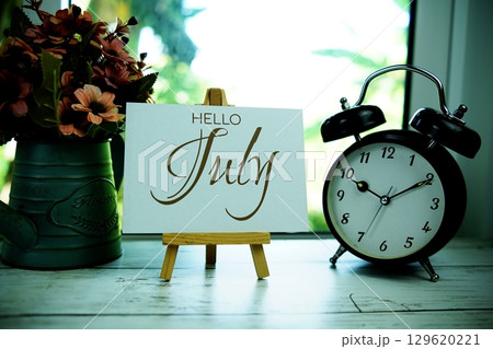 Hello July text on paper card with easel and alarm clock in the window background Hello July text on paper card with easel and alarm clock in the window background 129620221