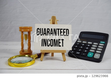 Guaranteed Income text with magnifying glass and calculator on wooden background Guaranteed Income text with magnifying glass and calculator on wooden background 129623966