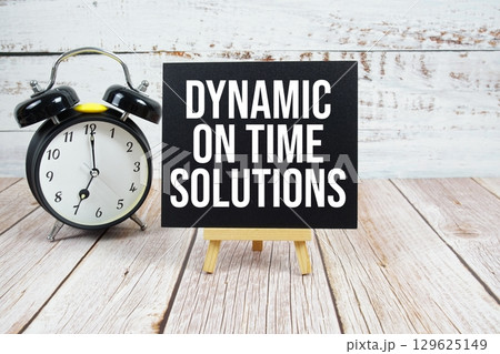 Dynamic On Time Solutions text with easel and alarm clock on wooden background Dynamic On Time Solutions text with easel and alarm clock on wooden background 129625149