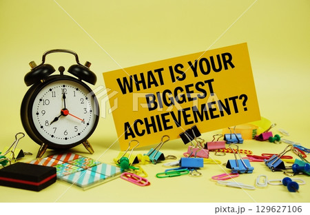 What is Your Biggest Achievement? text on paper card with alarm clock and stationery on yellow background 129627106