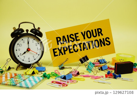 Raise Your Expectations text on paper card with stationery and alarm clock on yellow background Raise Your Expectations text on paper card with stationery and alarm clock on yellow background 129627108