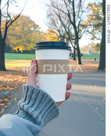 Woman Holding White Coffee Cup on a Sidewalk with Colorful Autumn Trees Background 129627988