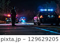 A police officer stands near patrol cars on a misty street at night, with blue and red lights reflecting on the wet road, creating a tense atmosphere of a potential crime scene 129629205