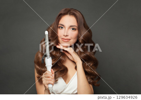 Lovely young model woman with long curly wavy hairstyle showing hot curling tongs iron 129634926