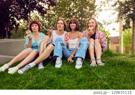 Group of young friends sitting on grass enjoying donuts in relaxed summer atmosphere Group of young friends sitting on grass enjoying donuts in relaxed summer atmosphere 129635118