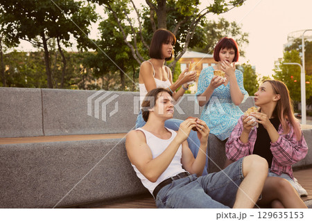 Friends relaxing and eating burgers and wraps on city bench 129635153