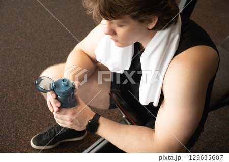 Man sitting in gym holding water bottle and towel on his neck. Moment of relaxation and refreshment after workout. Man sitting in gym holding water bottle and towel on his neck. Moment of relaxation and refreshment after workout. 129635607