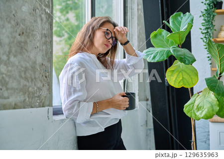 Mature tired, stressed woman standing near window Mature tired, stressed woman standing near window 129635963