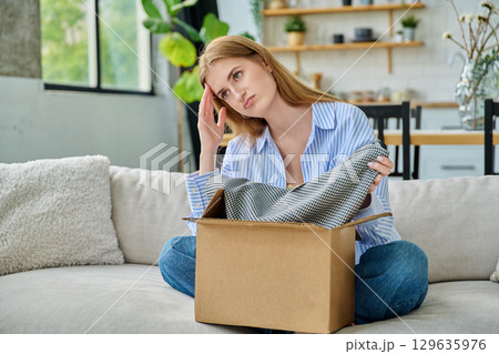 Sad young woman unpacking online purchase, looking at new clothes Sad young woman unpacking online purchase, looking at new clothes 129635976