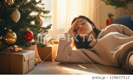 Young woman lying by a Christmas tree in sweatpants, surrounded by ornaments, golden hour lighting 129637512