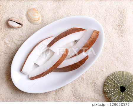 Fresh coconut meat, in oval white bowl, on beach sand with shells. Opaque white flesh of a fresh and young coconut, cut into strips, and ready to eat. Seed of the palm tree Cocos nucifera. Food photo 129638575
