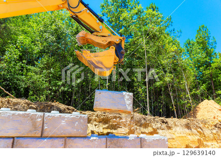 Heavy machinery lifts stone block at construction site surrounded by lush trees on works day 129639140