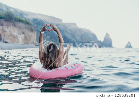 Woman, sea, relaxation. Young woman floats on pink donut in ocean making heart hands, enjoying summer vacation. 129639201