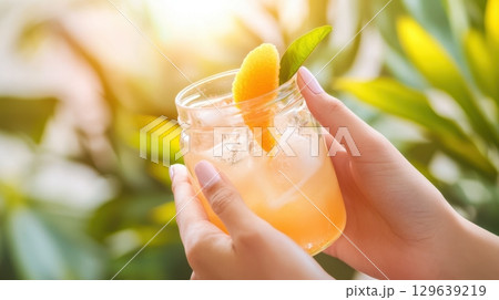 Female hands holding jam jar with peach jargarita cocktail, ice and orange peel on outdoor background with sunset light and greenery 129639219