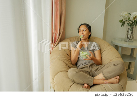 Relaxing Woman Happily Enjoys a Delicious Snack in a Cozy Living Room Space Relaxing Woman Happily Enjoys a Delicious Snack in a Cozy Living Room Space 129639256