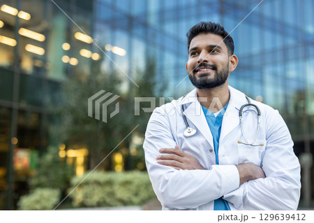 A confident doctor in a white coat smiling while standing outside near a modern building. A confident doctor in a white coat smiling while standing outside near a modern building. 129639412