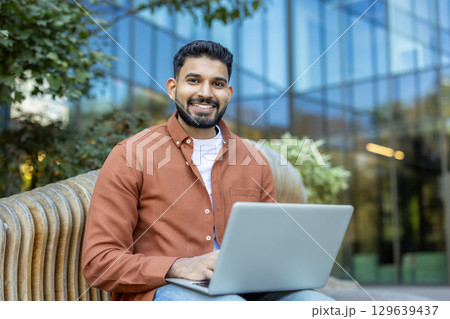A cheerful man in a casual outfit works on a laptop while seated outdoors, with a modern glass building and greenery in the background, capturing a blend of technology and relaxation. A cheerful man in a casual outfit works on a laptop while seated outdoors, with a modern glass building and greenery in the background, capturing a blend of technology and relaxation. 129639437