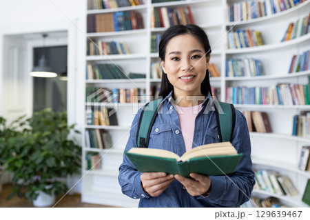 A young woman is smiling reading a book in a bright library setting. She carries a backpack, symbolizing education and knowledge, and the background includes rows of bookshelves and indoor plants. 129639467