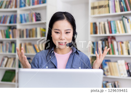 A young woman wearing a headset talking to someone via video call in a library; expressive gestures convey her active participation and interaction during the session. 129639481