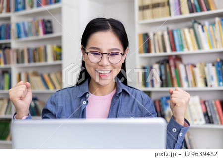 A happy young woman in glasses joyfully celebrates after accomplishing work on her laptop in a library, surrounded by bookshelves, reflecting achievement, success, and motivation. 129639482