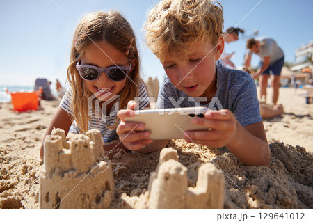 Children building sandcastles on the beach while capturing memories on a smartphone 129641012