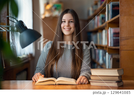 Female student sitting at the desk, reading a book and smiling. Brunette girl with long hair enjoys studying time at the library, getting education with pleasure Female student sitting at the desk, reading a book and smiling. Brunette girl with long hair enjoys studying time at the library, getting education with pleasure 129641085
