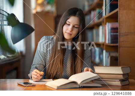 Concentrated young female student sitting at the table, reading and writing something for her studying. Young brunette girl working at the library, doing her hometasks 129641086