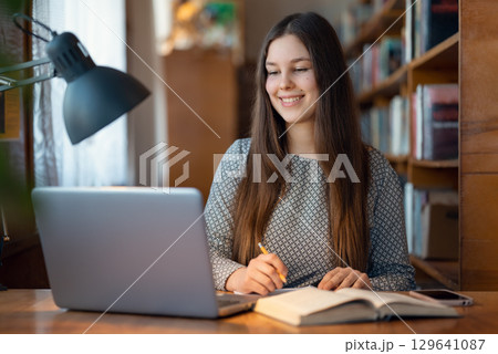 Cute young student girl sitting at a table with laptop in front of her, writing something in the notebook. Concept of education, studying in a library, modern students Cute young student girl sitting at a table with laptop in front of her, writing something in the notebook. Concept of education, studying in a library, modern students 129641087