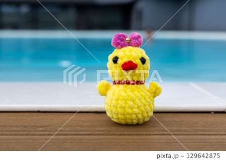 A cute, handmade crochet chick wearing a pink bow sits by a pool. Summertime vacation. A funky chicken toy against the blue water in a swimming pool. A cute, handmade crochet chick wearing a pink bow sits by a pool. Summertime vacation. A funky chicken toy against the blue water in a swimming pool. 129642875