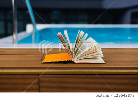 An open book sits peacefully near a pool offering a moment of relaxation A paper book rests on a wooden surface near a swimming pool Leisurely reading An open book sits peacefully near a pool offering a moment of relaxation A paper book rests on a wooden surface near a swimming pool Leisurely reading 129642878