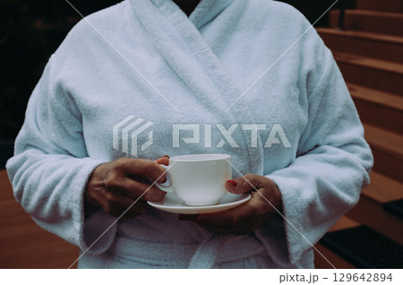 A relaxed woman wearing a white bathrobe holds a cup of coffee enjoying peaceful moment. A person in a soft bathrobe holding a mug, relaxing in hotel. 129642894