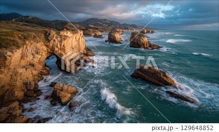 Remote windswept coastline with dramatic rock arch, rugged cliffs, ocean waves, moody sky 129648852