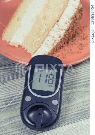 Glucometer with result of sugar level and cake with layers and chocolate. Nutrition during diabetes Glucometer with result of sugar level and cake with layers and chocolate. Nutrition during diabetes 129655454